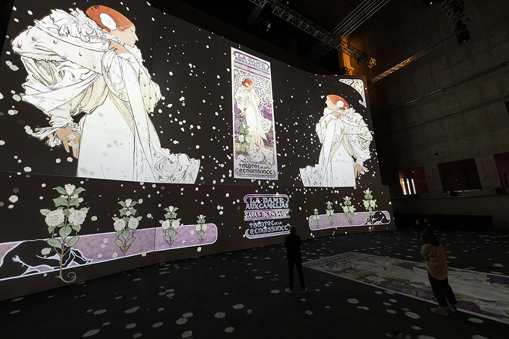 A dark room with large screens brightly projecting a woman in a white dress surrounded by flowers onto the walls and floor to create an immersive experience