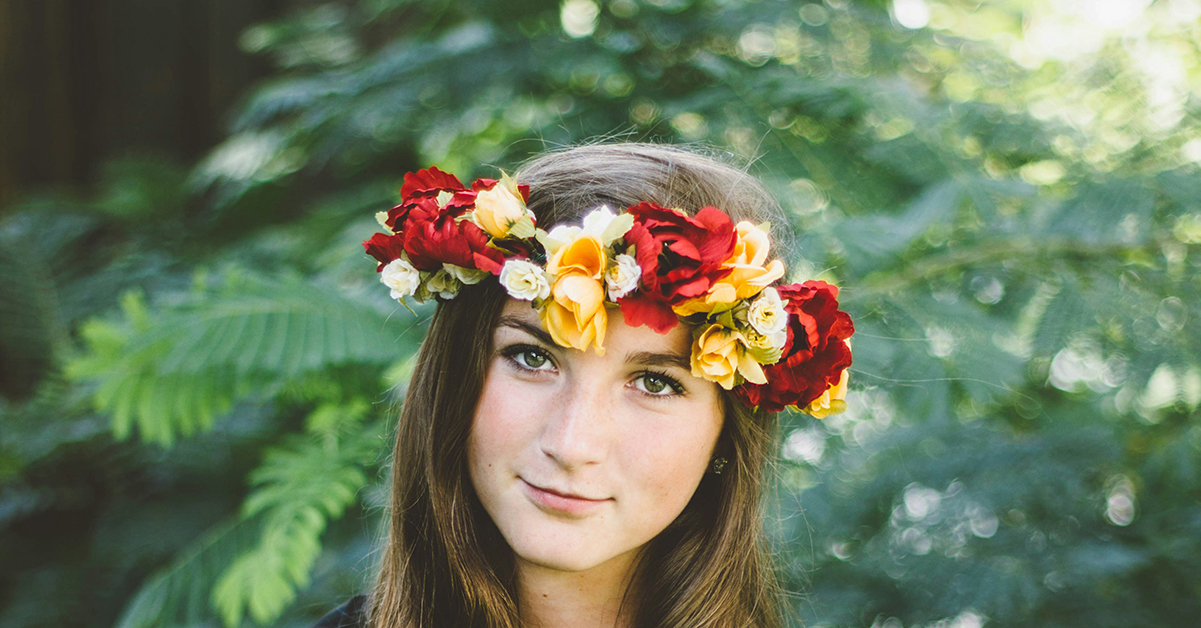 A young woman with long brown hair wearing a flower crown with green leaves and ferns behind her