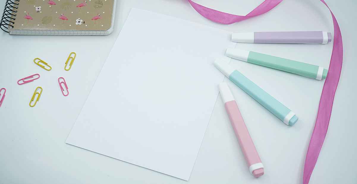 White notecard with pastel markers, ribbon, paperclips and a notebook on a white table