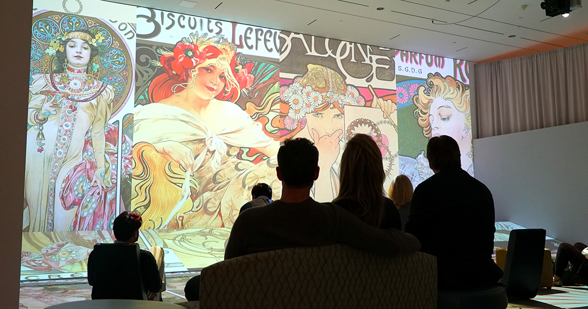 A group of people seated in a gallery looking at a brightly colored immersive featuring the work of Alphonse Mucha